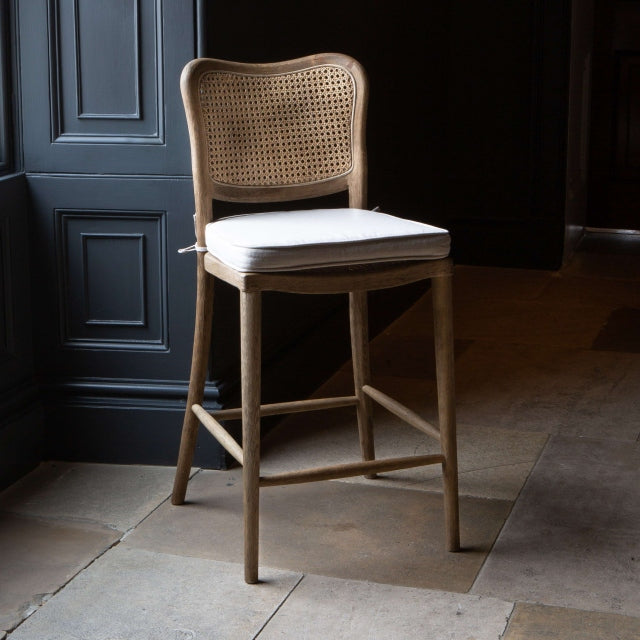 A barstool with a natural linen upholstered seat pad and a wood finish. The stool has a rattan backrest and is placed against a dark background.