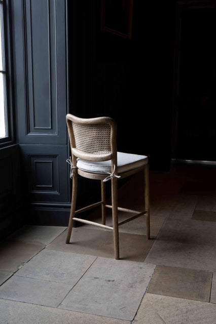 Back view of A barstool with a natural linen upholstered seat pad and a wood finish. The stool has a rattan backrest and is placed against a dark background.