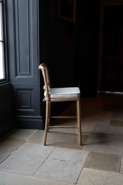 Side view of A barstool with a natural linen upholstered seat pad and a wood finish. The stool has a rattan backrest and is placed against a dark background.