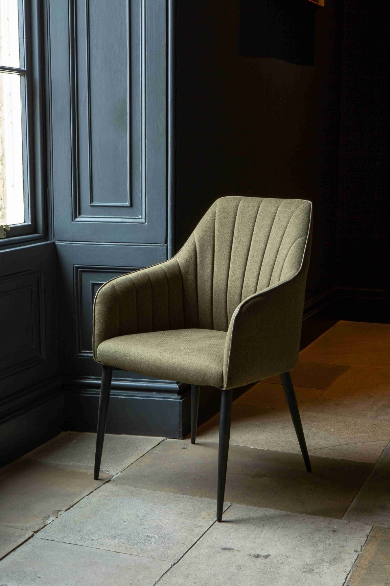 A green upholstered Brent dining chair with a sleek backrest and black metal legs facing a window positioned against a dark background.
