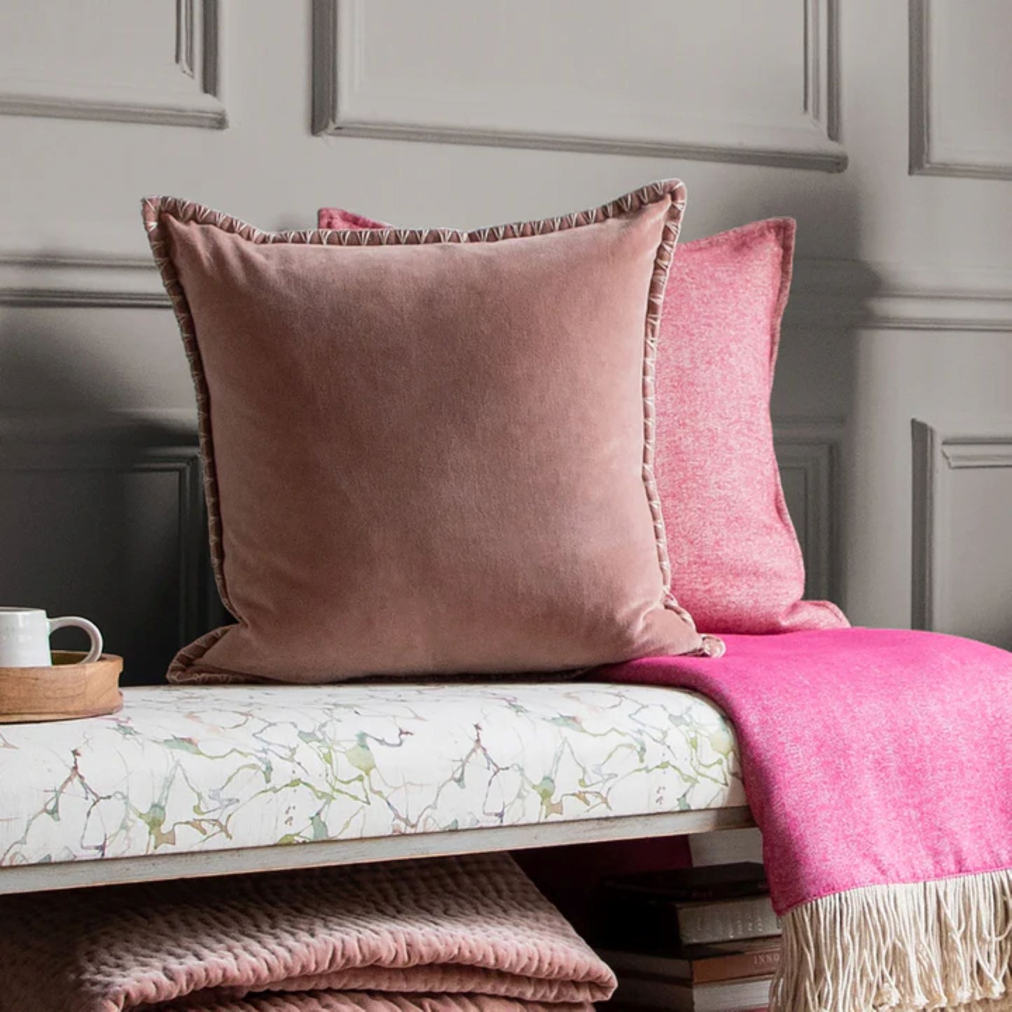 A dusky pink velvet cushion with a stitch detail edge bench seat, surrounded by other cushions and a pink throw.