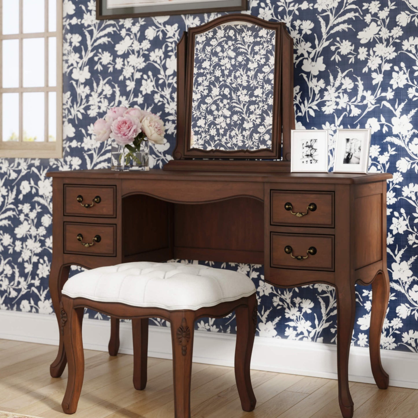 An elegant brown wooden dressing table with four drawers and a stool with a white cushion, positioned against a wall with floral wallpaper.