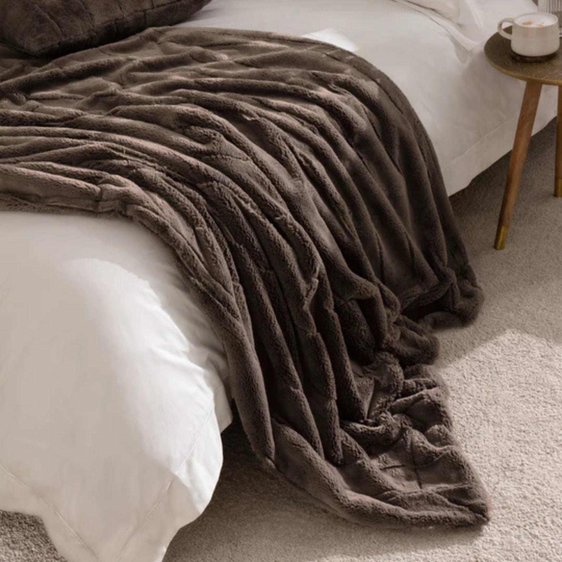 Brown faux fur throw draped acrross a bed with white linen and a small round wood side table with a cup on it