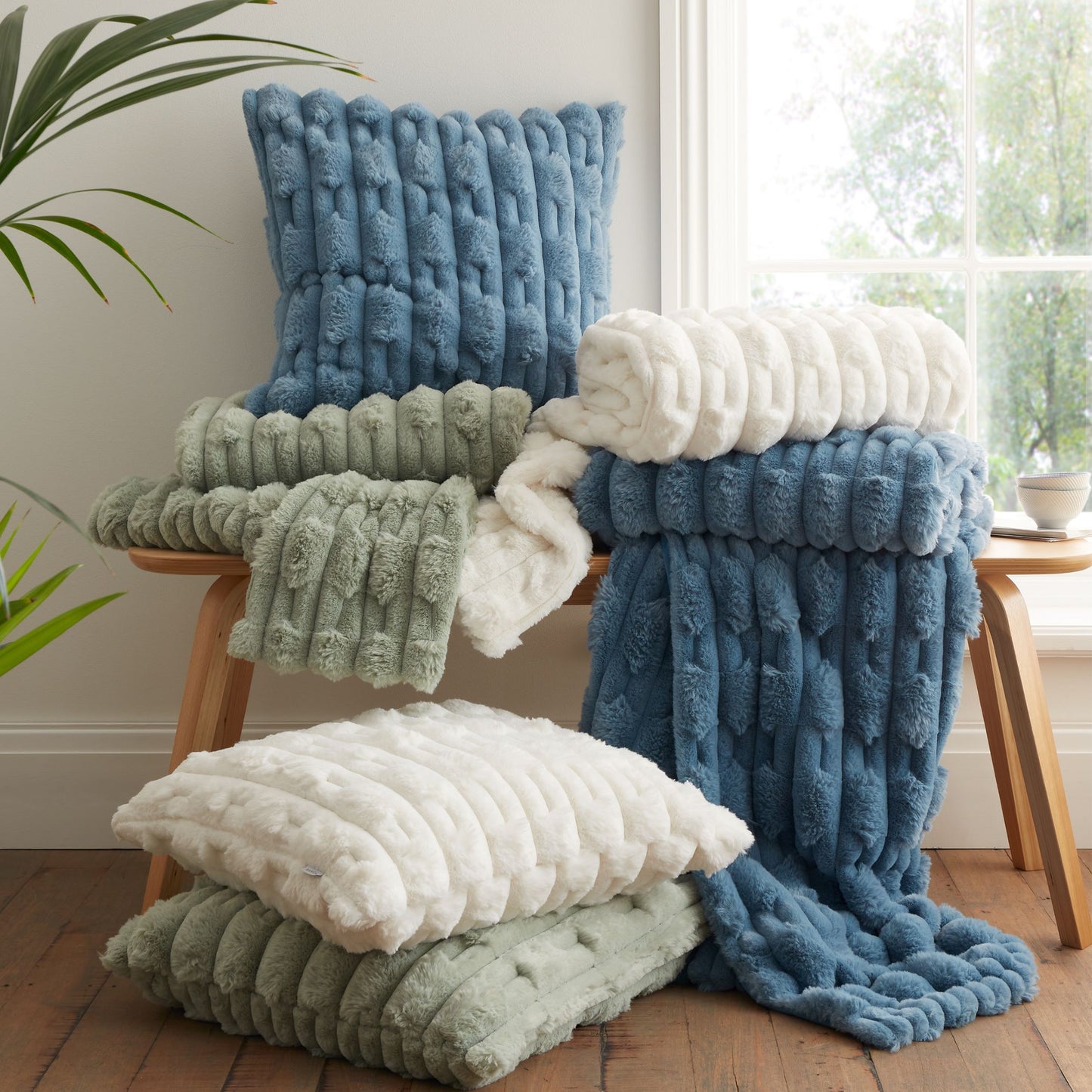 Faux fur blue, cream and green cushions and throws draped on a wooden bench in front of a window with 2 cushions on the floor.