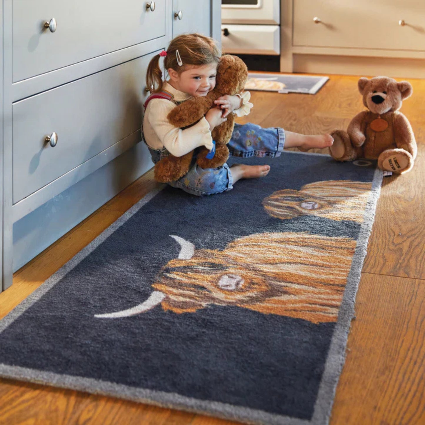 Child playing with teddy bears on a rug with highland cow design in a kitchen.