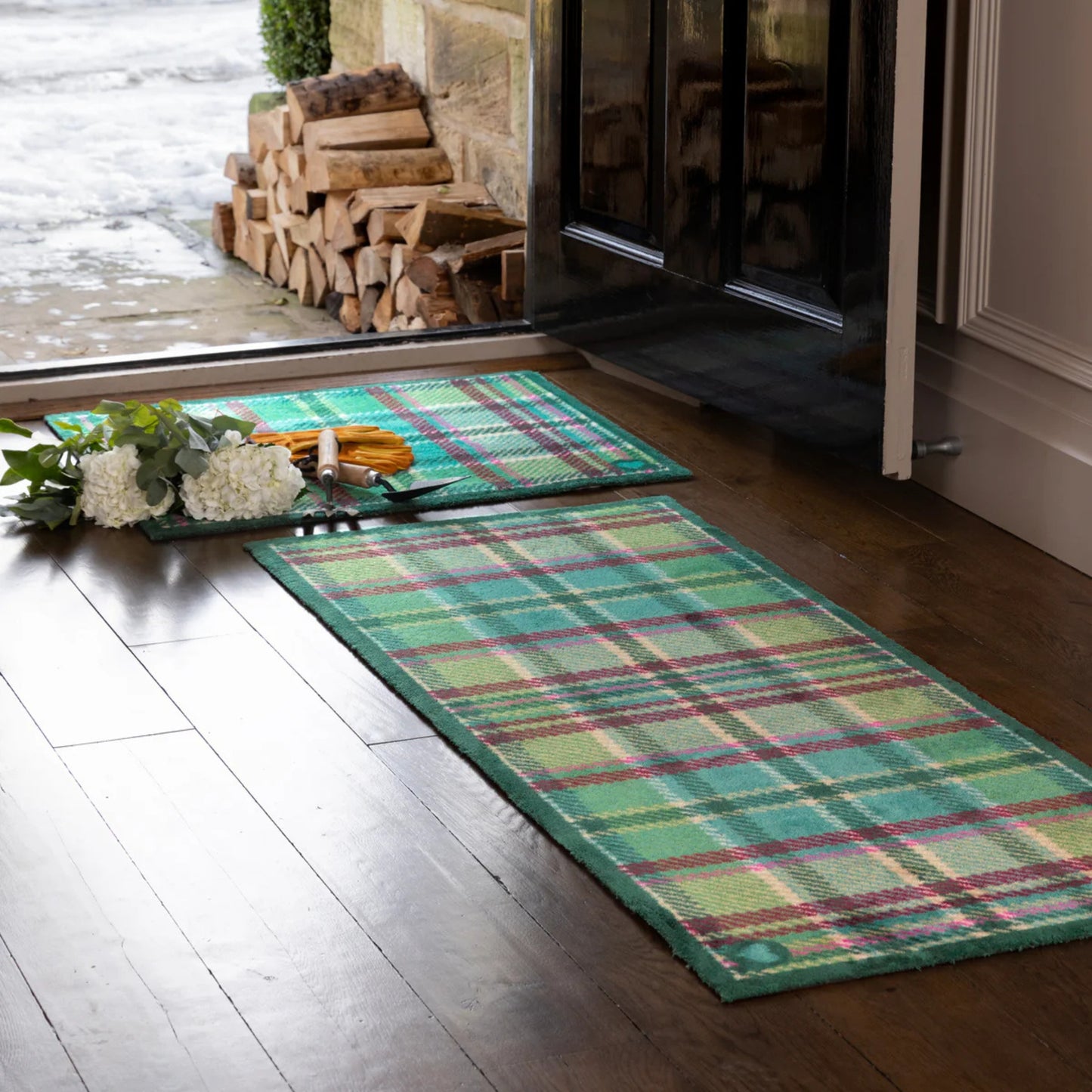 Green plaid rug on a wooden floor with a glass door and stacked firewood in the background.