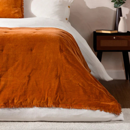 Orange bedspread accross a bed dressed in white linen with a dark wood bedside table with a book and two vases sitting on top.