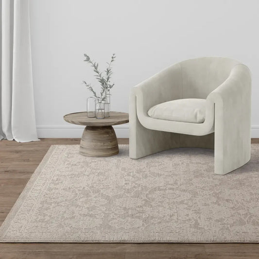 A beige Turkish rug placed on the floor under a modern armchair, with a round wooden side table and a vase with flowers on top.