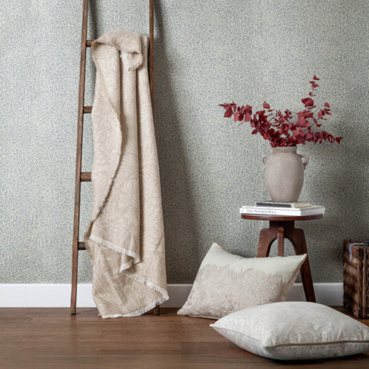 Beige blanket draped over a wooden ladder against a textured wall with pillows and a vase of flowers on a small table.
