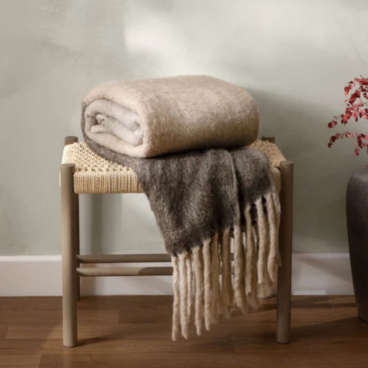 Folded fringed throw blanket on a woven stool against a neutral wall.