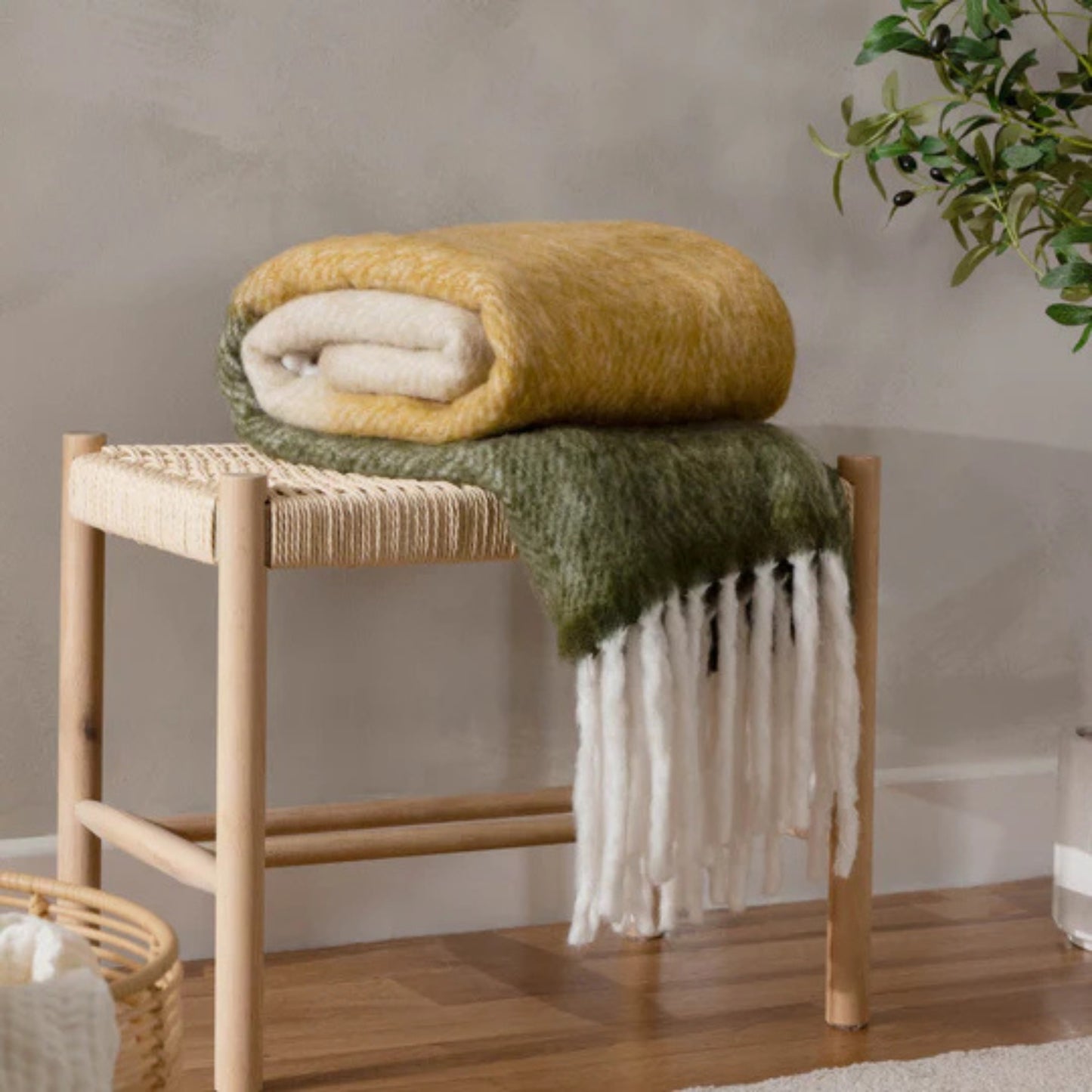 Woven stool with folded blanket on a wooden floor, plant in the corner