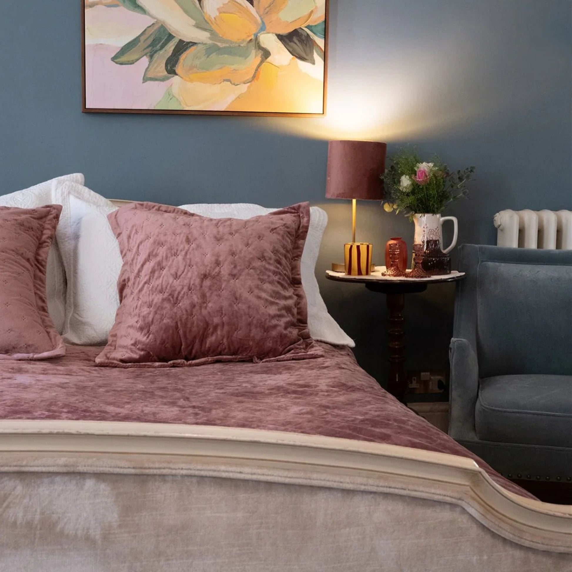 Bedroom setting with art hanging over a bed dressed in pink velvet bedspread and cushion with small side table and lamp next to blue accent chair