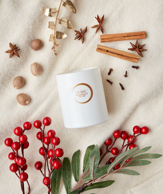 A white tumbler candle with a label, surrounded by decorative elements like cinnamon sticks, cloves, red berries, and green leaves.