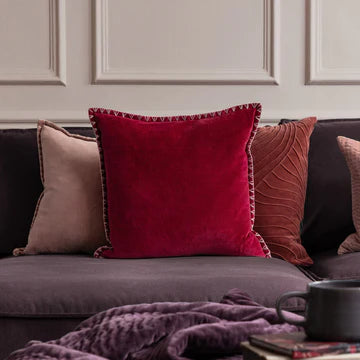 A red velvet cushion with a stitch detail edge on a grey sofa, surrounded by other cushions and a purple throw.