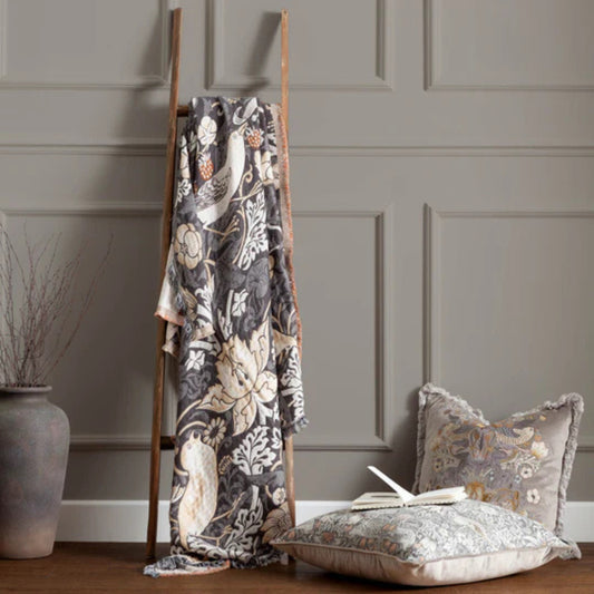 Patterned throw hanging on wooden ladder against a greige background with a stone vase and cushions next to it.