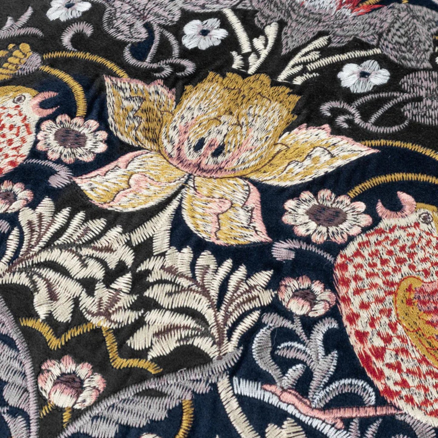 A square-shaped, indigo-colored velvet cushion close up detail with a floral and animal embroidery pattern, featuring a central bird and surrounding floral designs