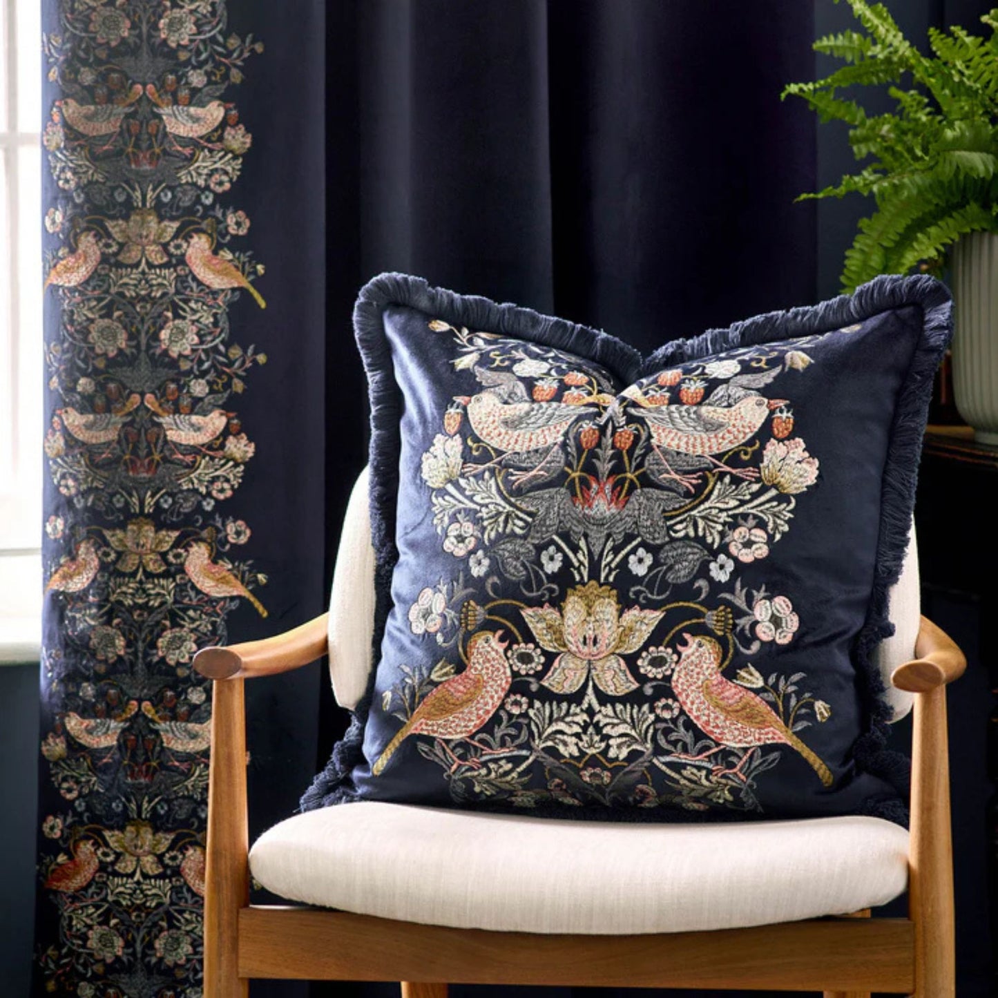 A square-shaped, indigo-colored velvet cushion with a floral and animal embroidery pattern, featuring a central bird and surrounding floral designs, finished with an accent fringed edging, sitting on a chair in a living room setting