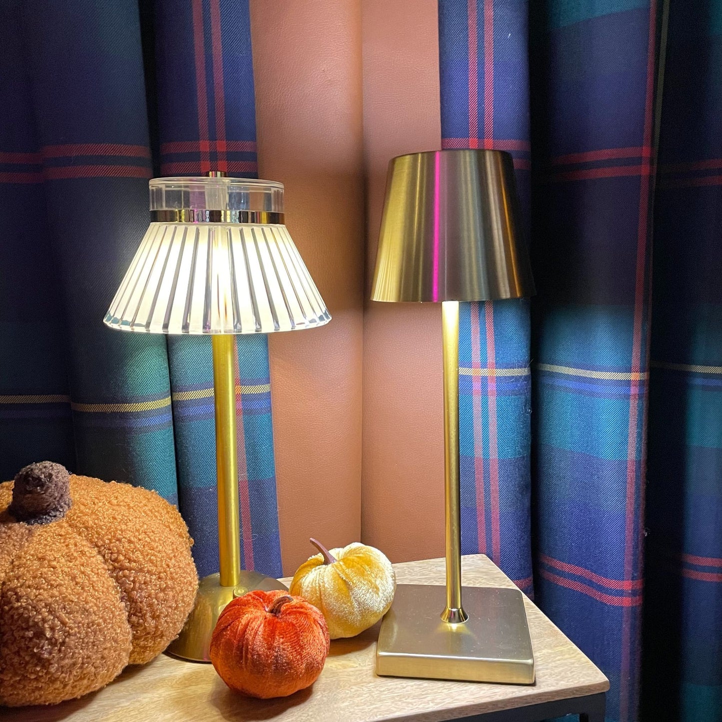 A portable antique gold rechargeable LED lamp with a conical shade, displayed with an pleated shade rechargeable lamp against a background of check navy curtains and decorative pumpkins and a pheasant design cushion.