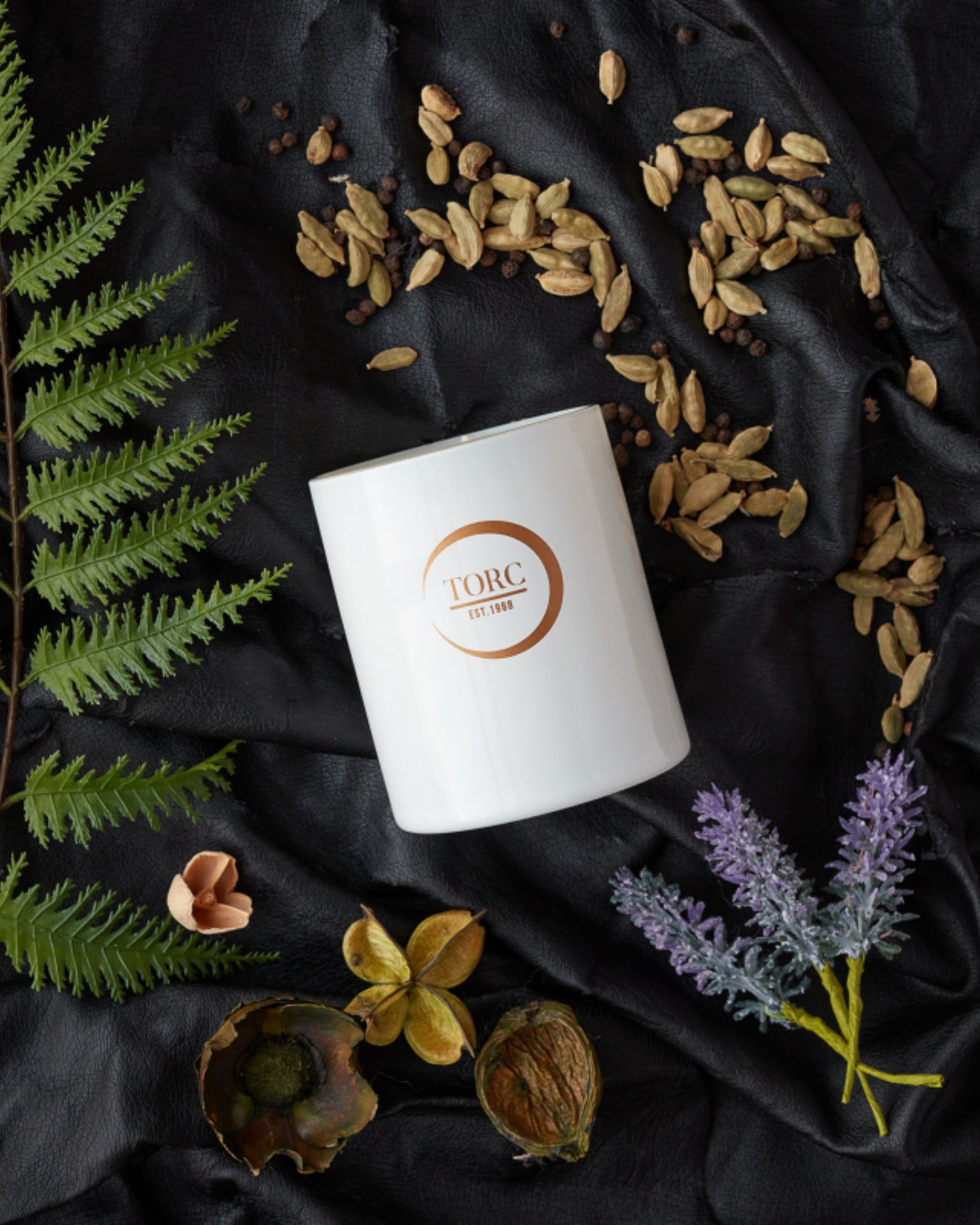 White candle with a logo on a black fabric background with dried leaves and flowers