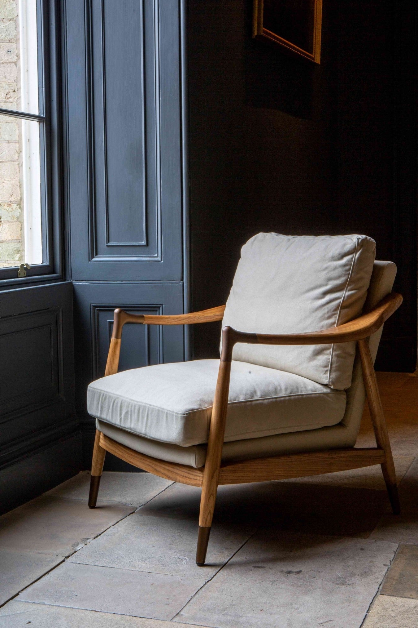 An elegant Archie Accent Chair with a solid ash timber construction and an upholstered ivory canvas seat and back cushion facing a window on a tiled floor with dark navy painted walls.