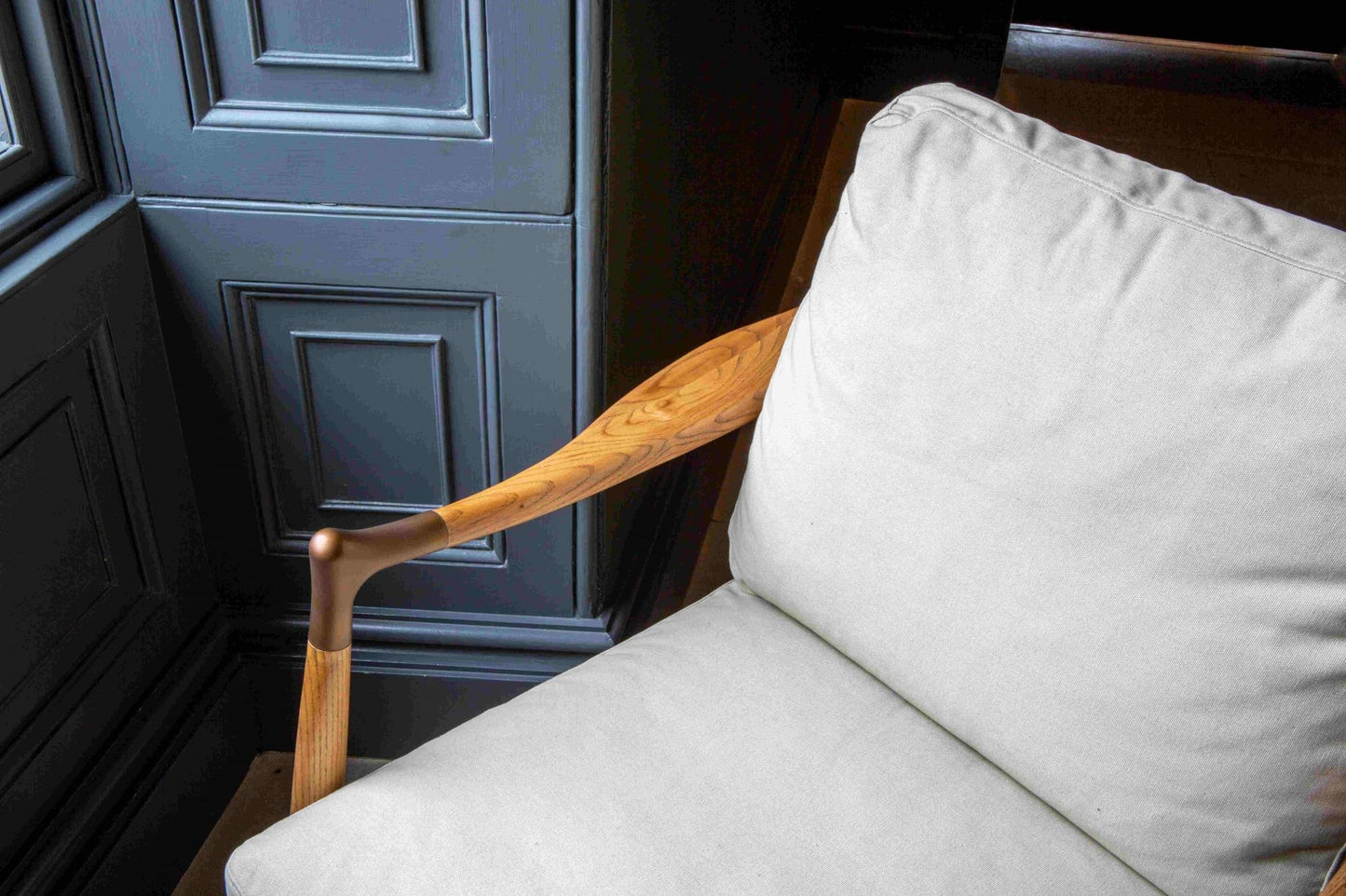 Detail close up of An elegant Archie Accent Chair with a solid ash timber construction and an upholstered ivory canvas seat and back cushion facing a window on a tiled floor with dark navy painted walls.