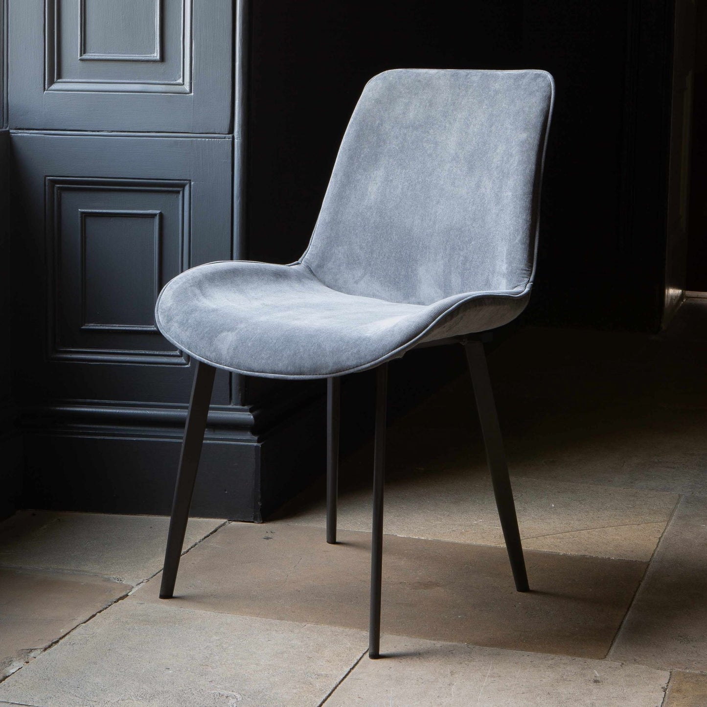A grey Eva dining chair with velvet upholstery and grey legs positioned in a room with a dark door in the background.