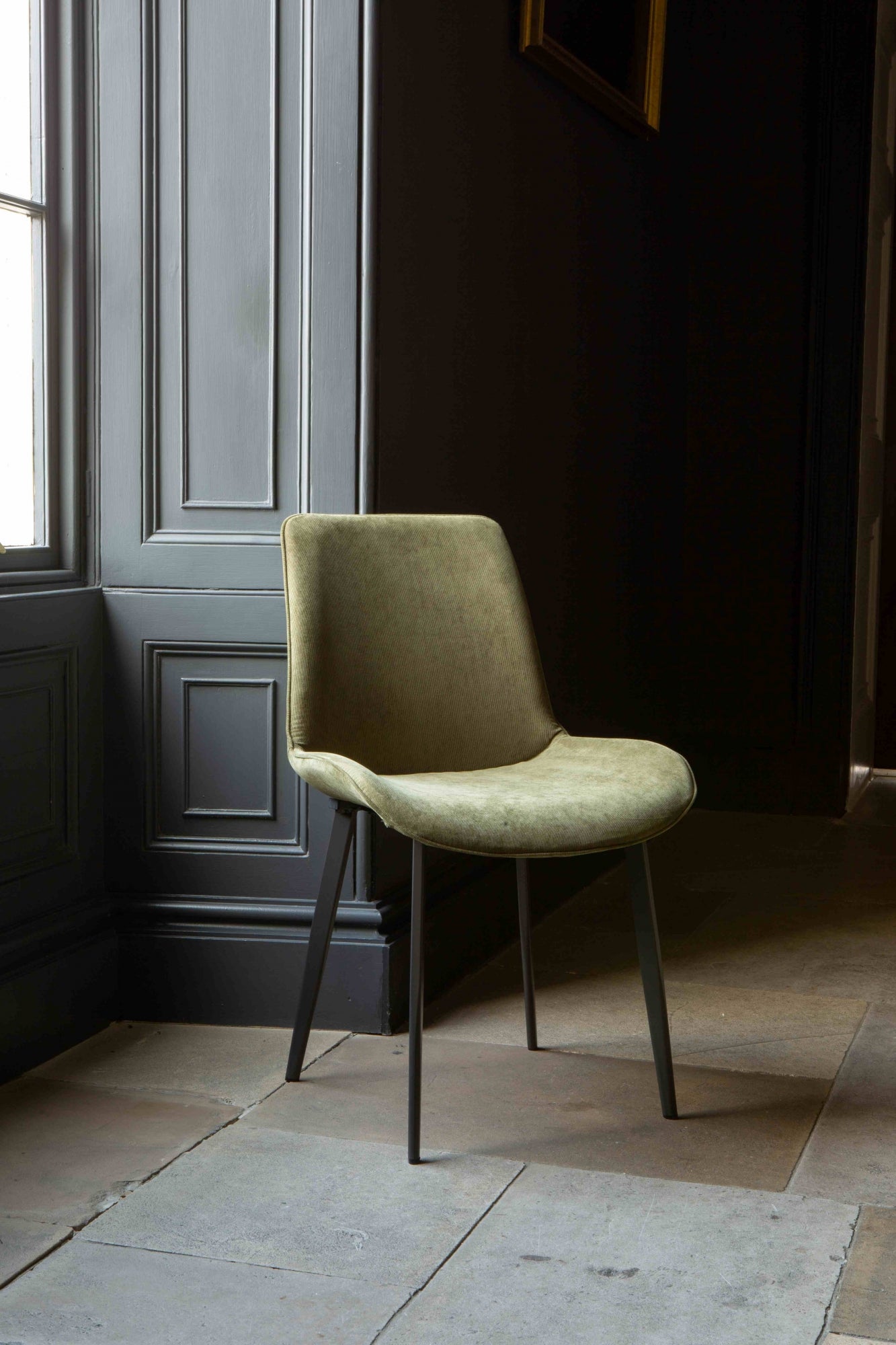 A green Eva dining chair with velvet upholstery and grey legs positioned in a room with a dark door in the background.