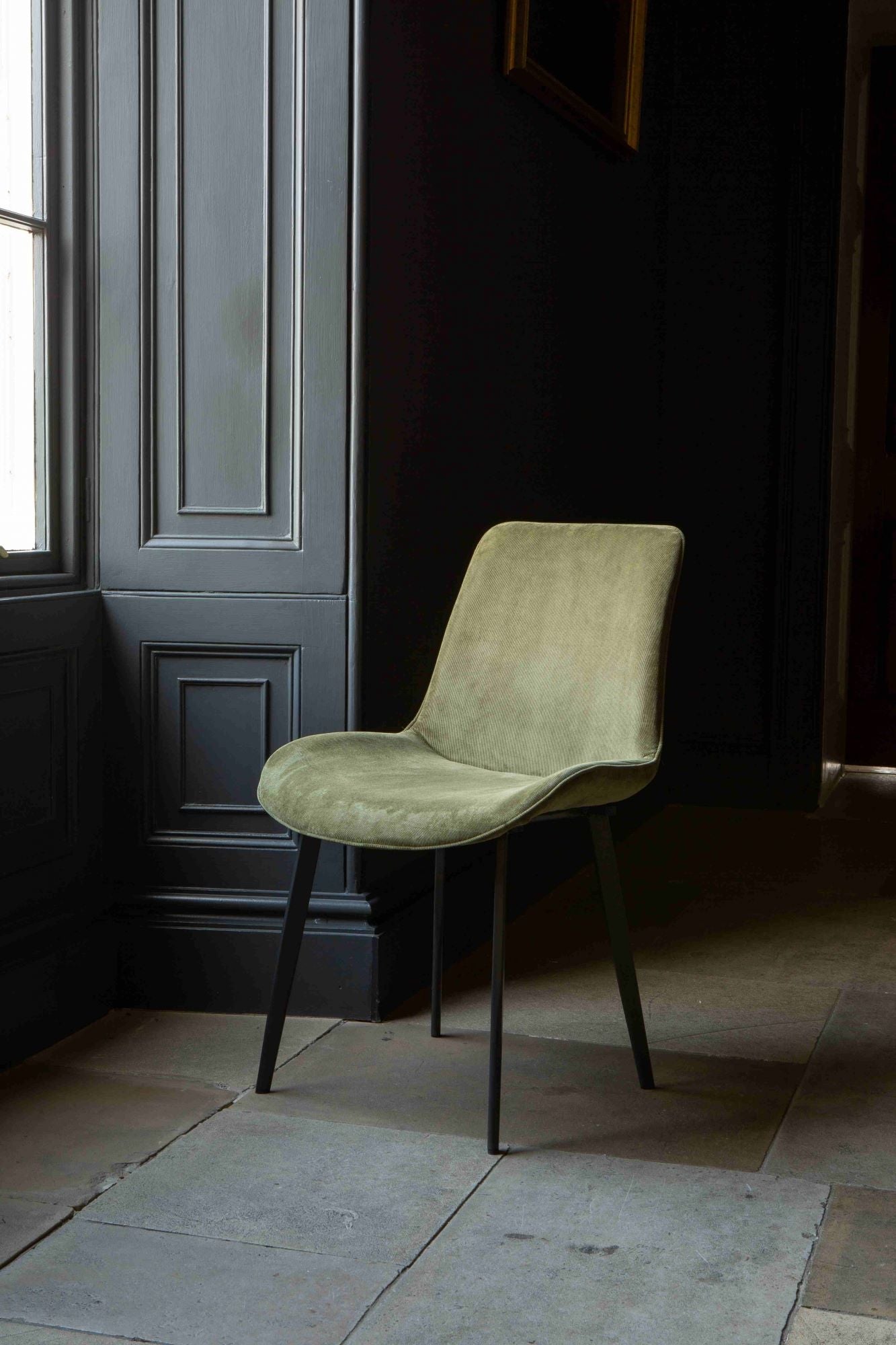 A green Eva dining chair with velvet upholstery and grey legs positioned in a room with a dark door in the background.