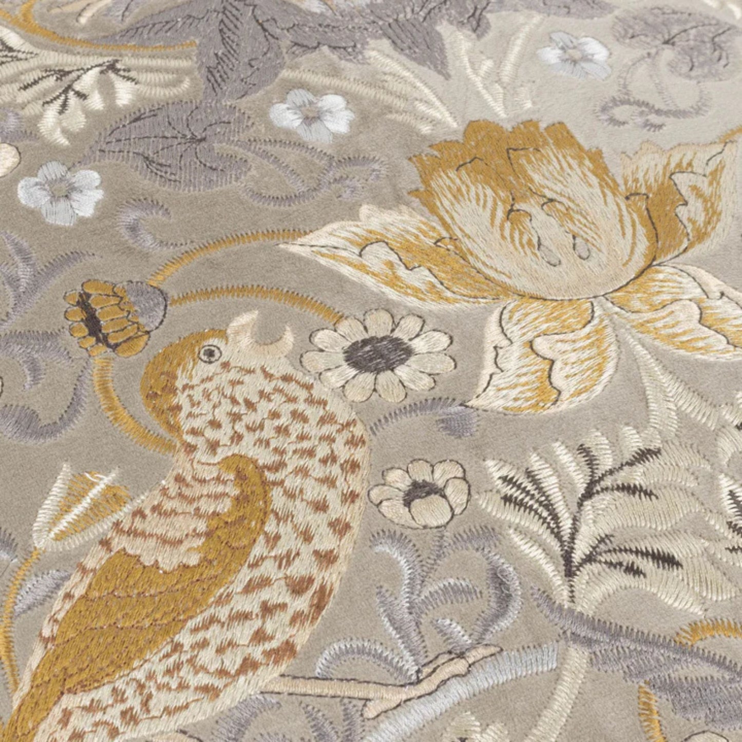 Detail close up of a biscuit coloured velvet cushion with a floral and animal embroidery pattern, featuring birds and surrounding floral designs, finished with an accent fringed edging.