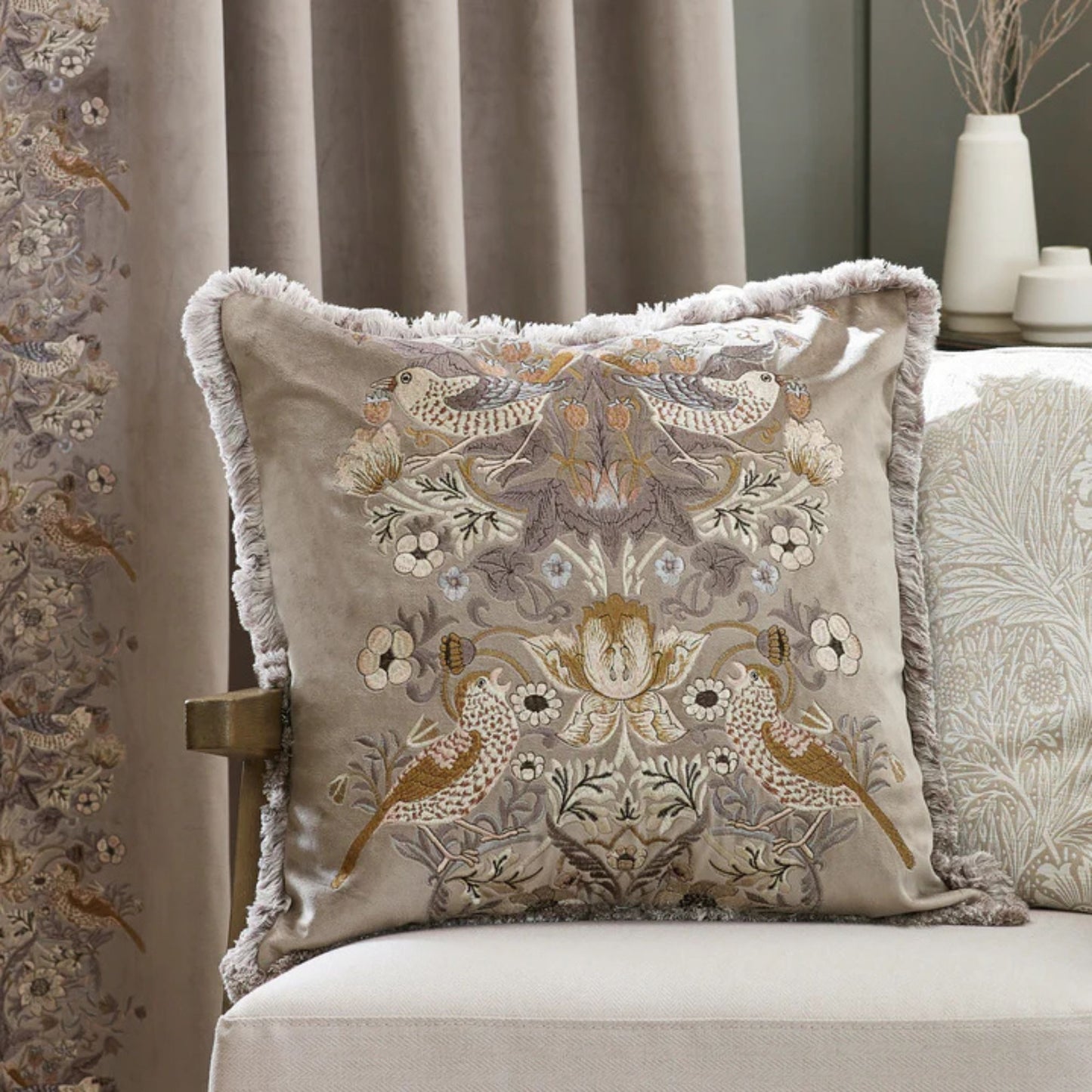 A square-shaped, biscuit coloured velvet cushion with a floral and animal embroidery pattern, featuring birds and surrounding floral designs, finished with an accent fringed edging, Sitting on a chair in a living room setting.