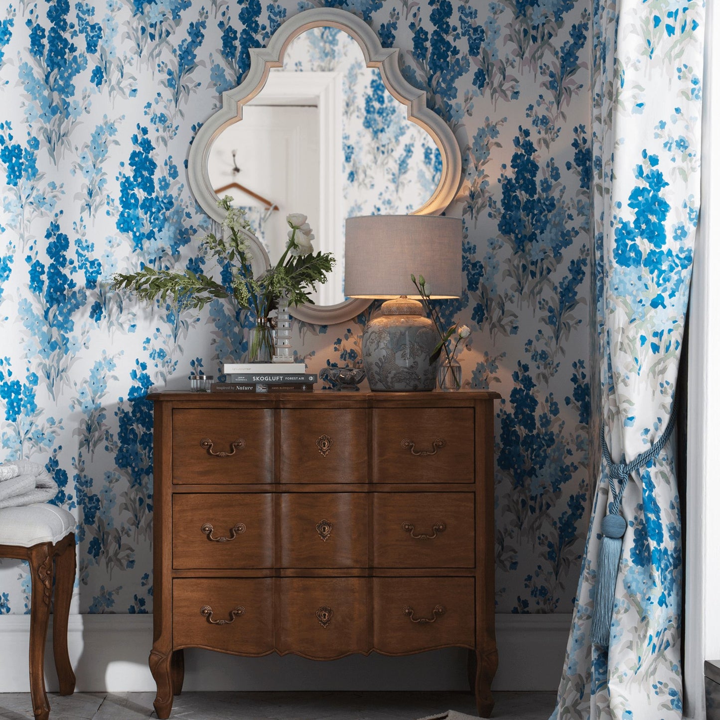 walnut 3 drawer chest styled ina bedroom featuring blue print wallpaper. Chest is styled with a lamp and vase of flowers with a decorative mirror hanging above it.