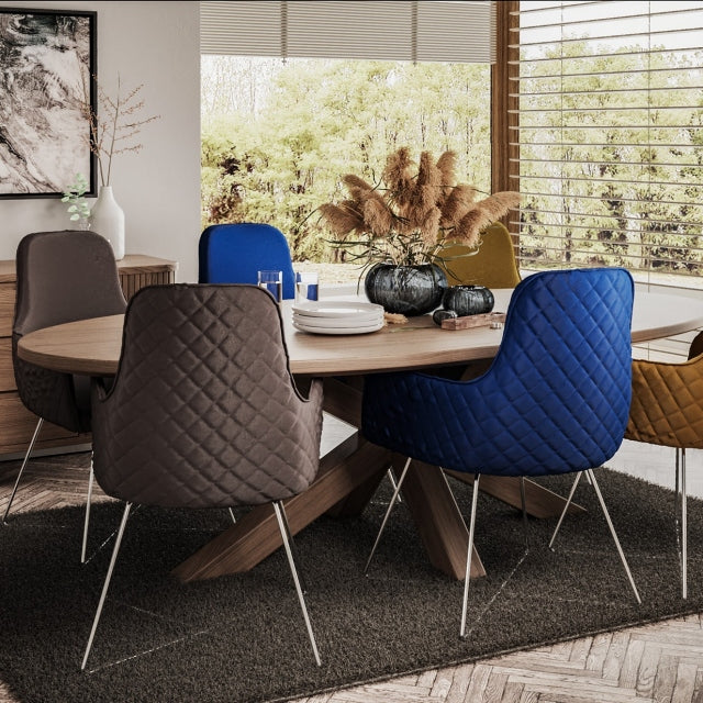 An oval-shaped solid wood oak dining table with double cross legs, displayed in a natural wood finish, with colourful velvet dining chairs  displayed on a grey rug n a dining room setting next to large windows.