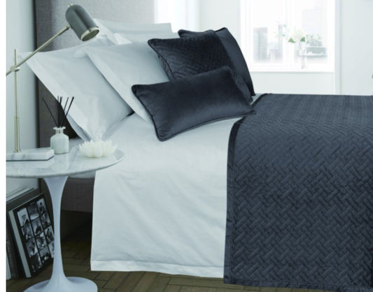 A navy and teal French velvet bedspread with hand-stitched quilting displayed on a bed in a bedroom setting, accompanied by cushions and a side table with a lamp.