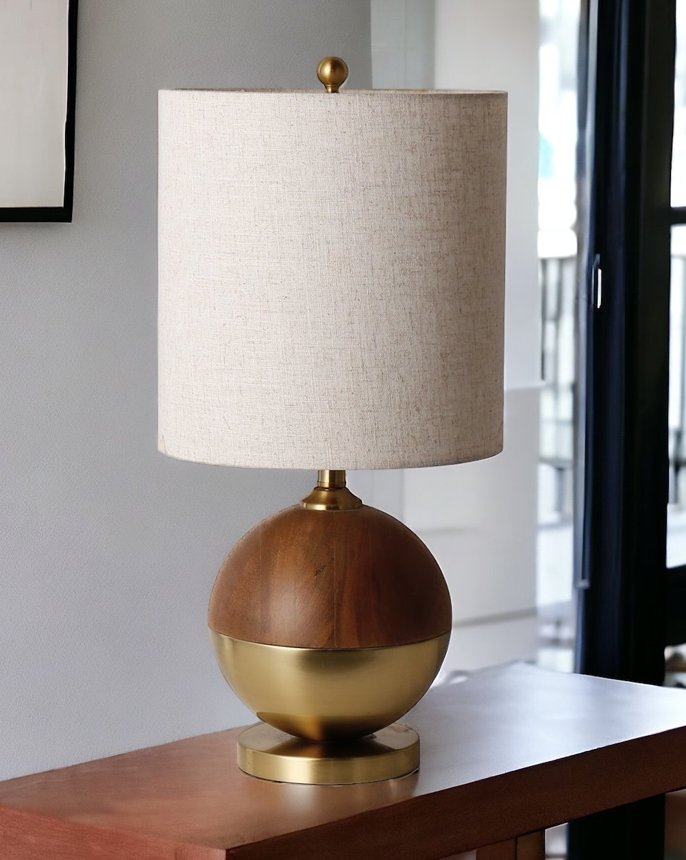 A modern table lamp with a large, rounded translucent amber glass base, a gold-finished metal stem and base, and a cylindrical off-white fabric drum shade, shown against a plain white background.