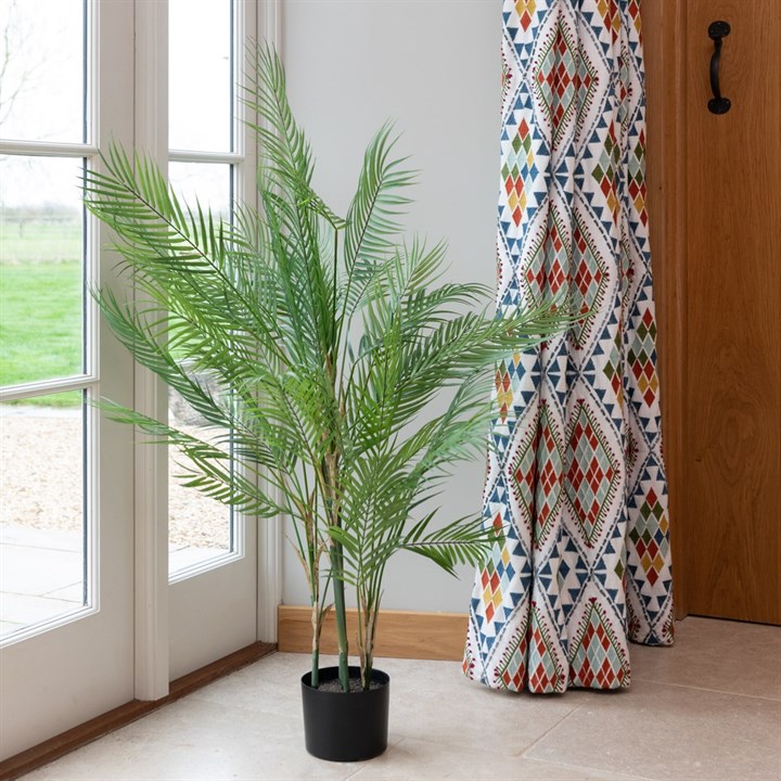 A faux areca palm plant with vibrant green leaves in a black pot sitting on the floor next to window with a colourful floor length curtain