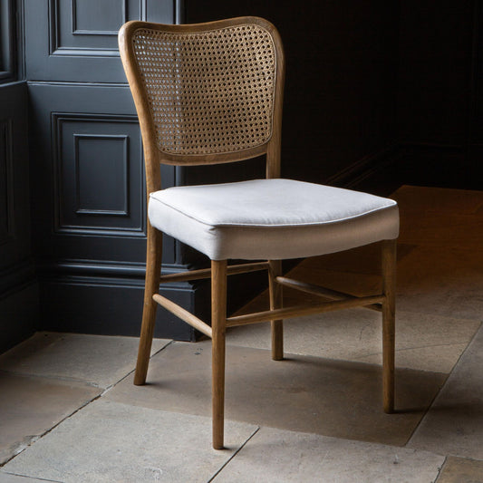 A dining chair with a natural linen upholstered seat pad and a wood finish. The back of the chair has a rattan weave design.