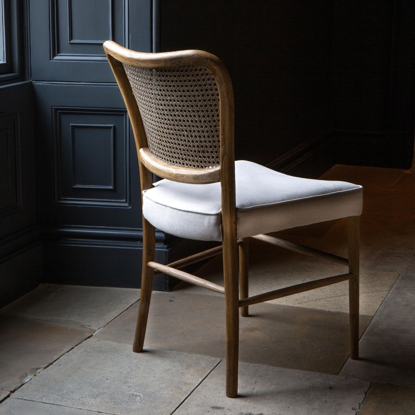 Back view of A dining chair with a natural linen upholstered seat pad and a wood finish. The back of the chair has a rattan weave design.