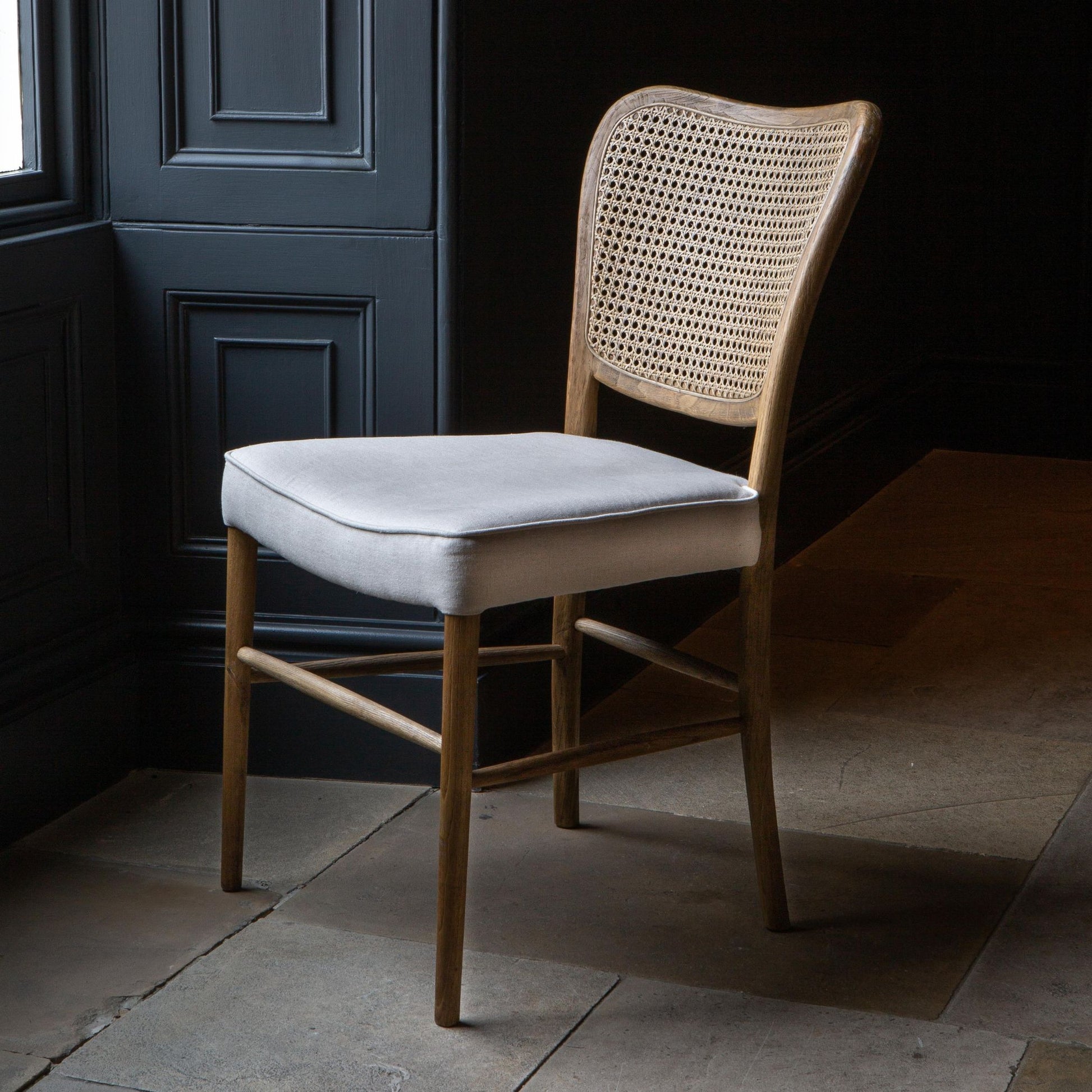 Front side view of A dining chair with a natural linen upholstered seat pad and a wood finish. The back of the chair has a rattan weave design.