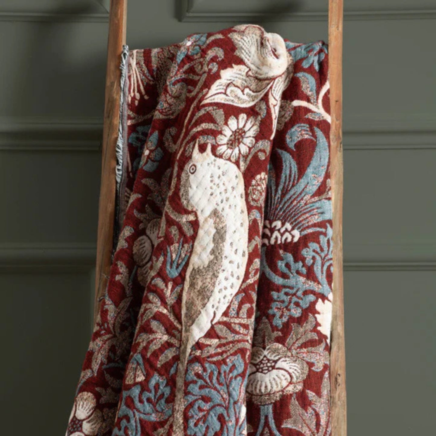 Decorative blanket with floral pattern draped over a wooden ladder against a gray wall.