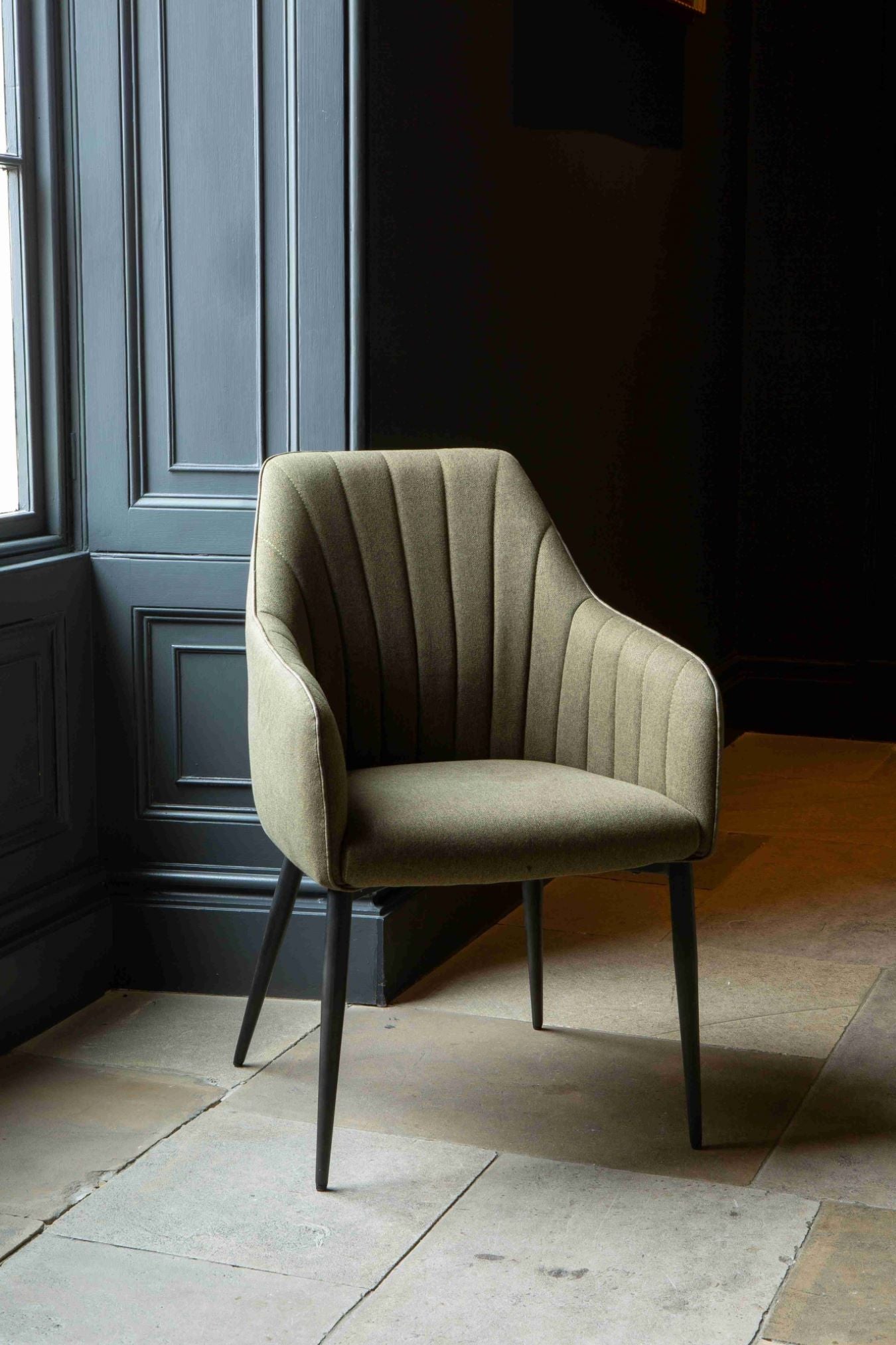 A green upholstered Brent dining chair with a sleek backrest and black metal legs positioned against a dark background.