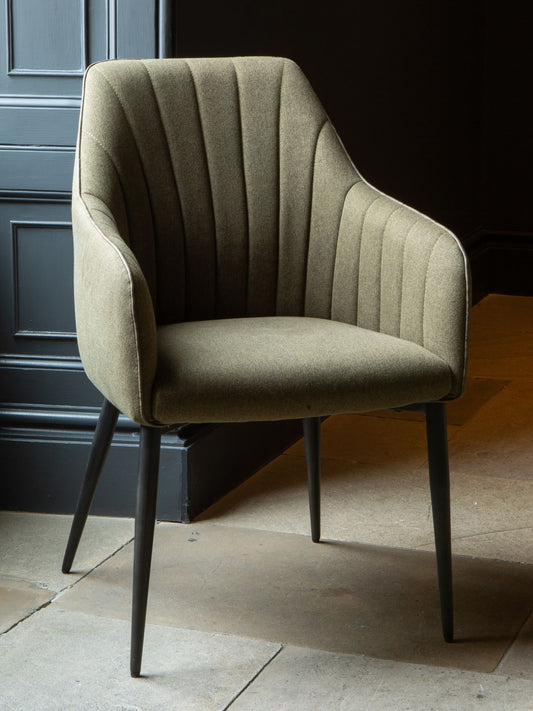 A green upholstered Brent dining chair with a sleek backrest and black metal legs positioned against a dark background.