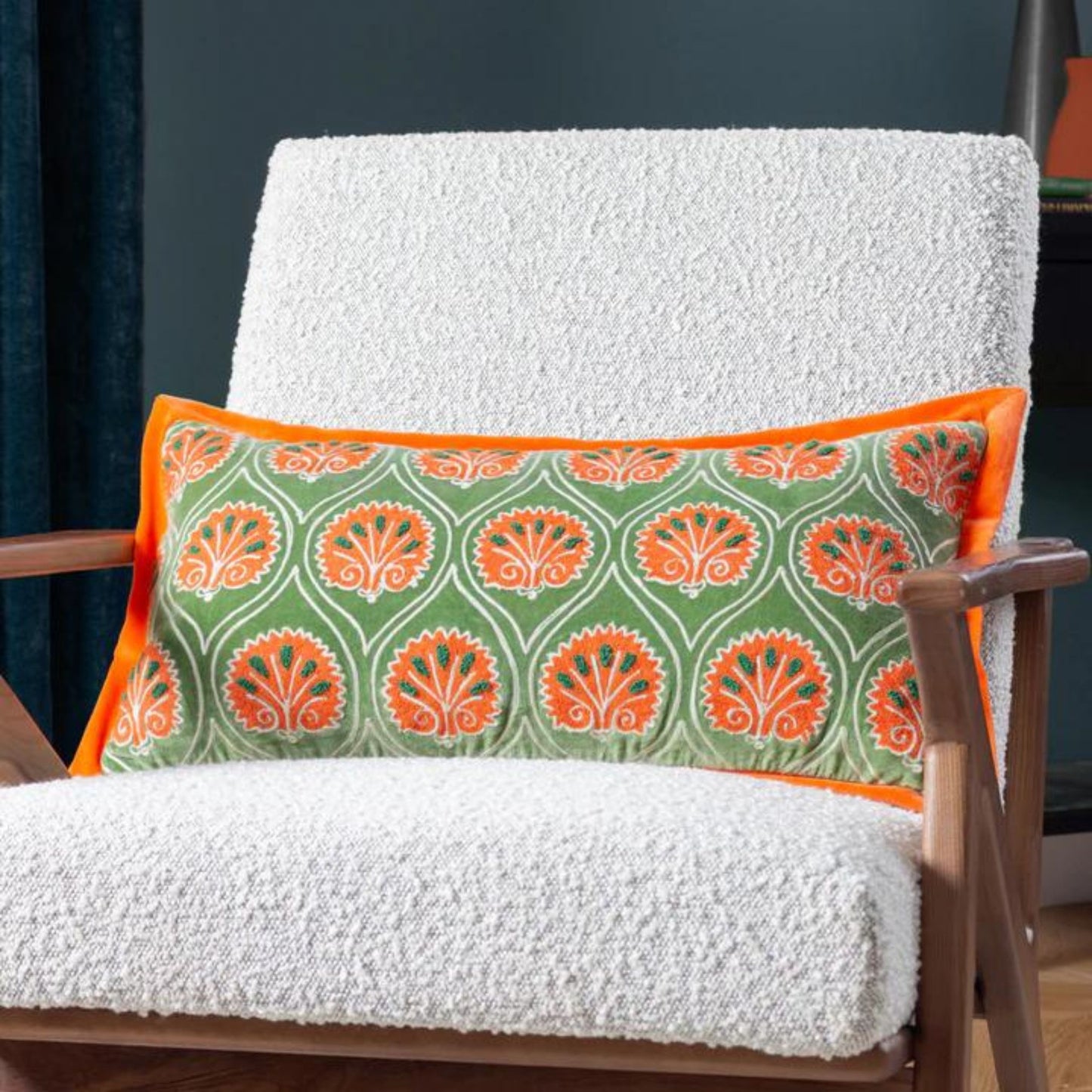 A rectangular orange and green embroidered cushion with a floral pattern, placed on a white sofa cushion on a wooden chair.