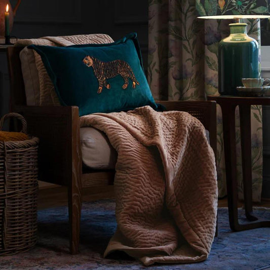 A soft coral velvet throw draped over the arm of a chair in a cozy room setting, with cushions and a lamp in the background.