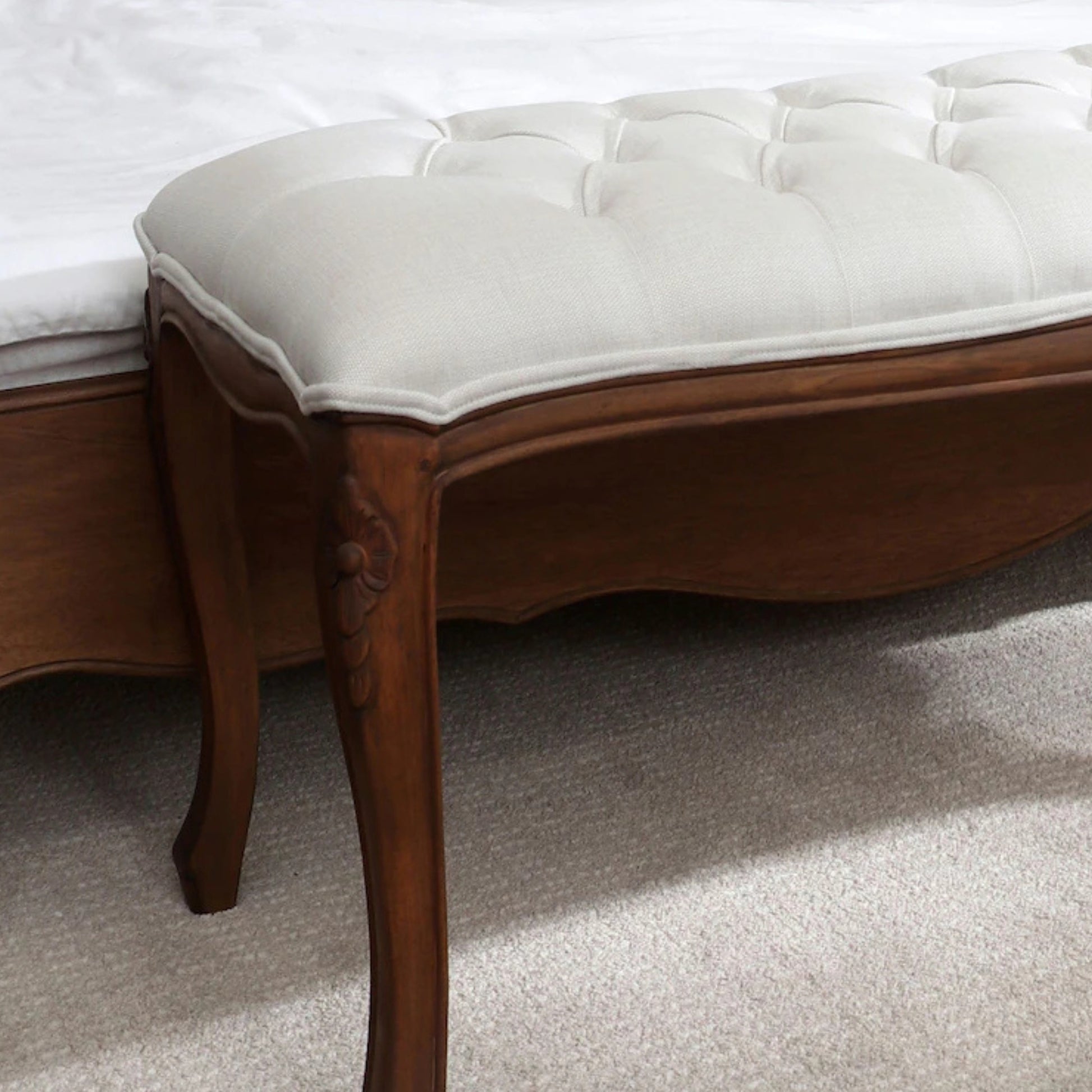Close-up of the front left leg and seat of an upholstered bench with a tufted backrest, showcasing a solid wood frame in an aged walnut finish and a beige upholstered top.