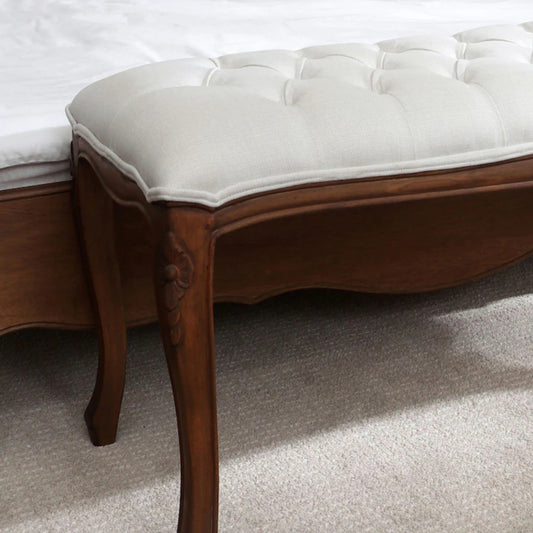 Close-up of the front left leg and seat of an upholstered bench with a tufted backrest, showcasing a solid wood frame in an aged walnut finish and a beige upholstered top.