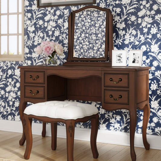 An elegant brown wooden dressing table with four drawers and a stool with a white cushion, positioned against a wall with floral wallpaper.