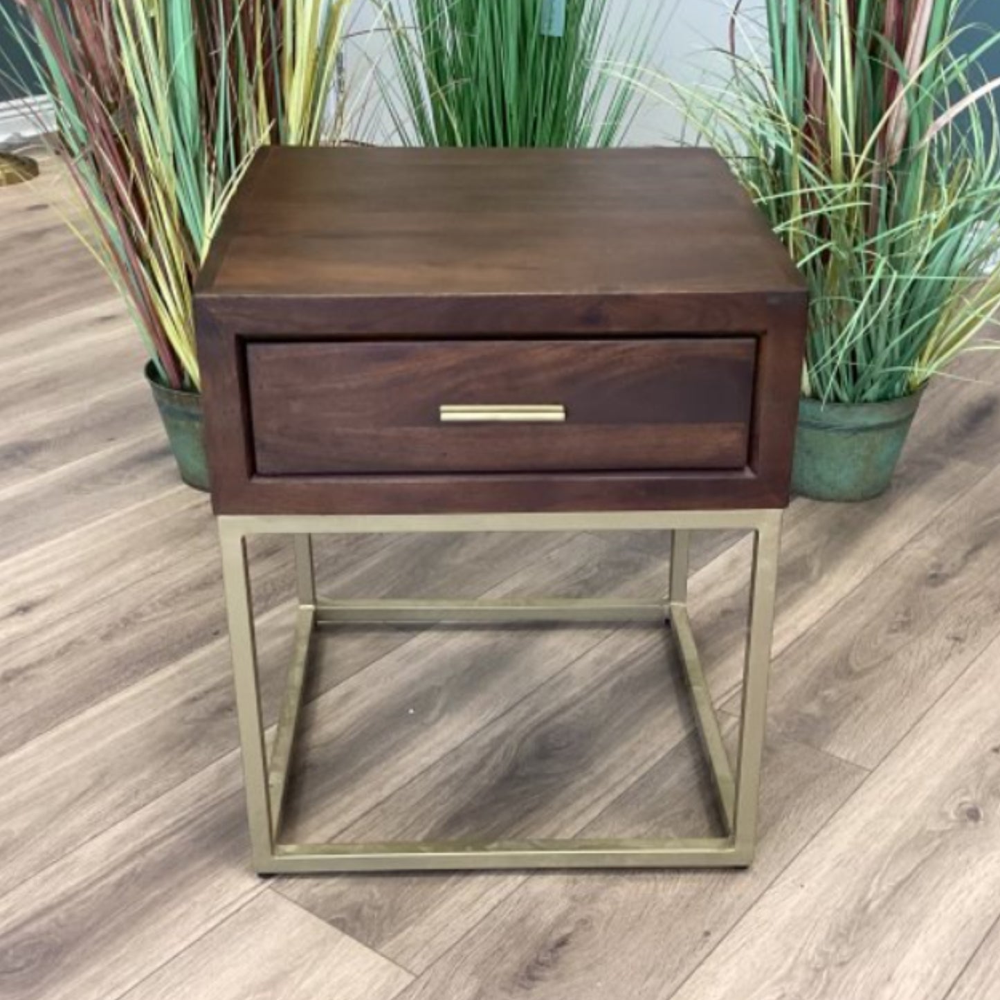 Wooden side table with a drawer and metal frame on a wooden floor with plants in the background