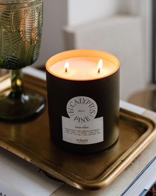 A black candle with two wicks labeled 'Eucalyptus & Pine' burning inside a transparent glass jar placed on a metal tray, with a blurred background.