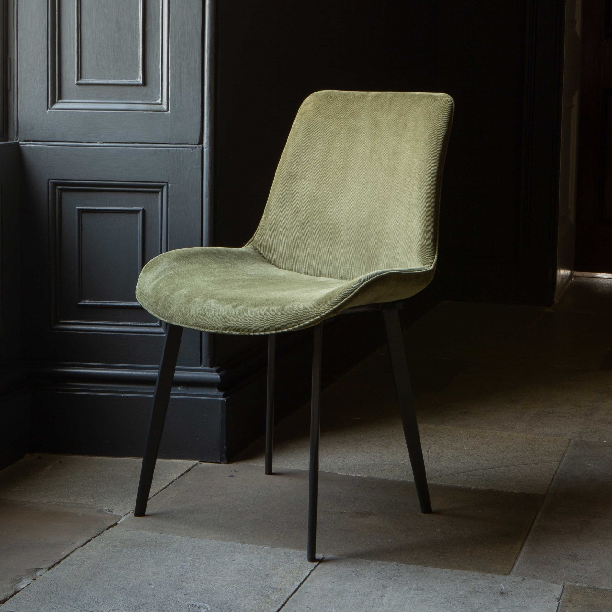 A green Eva dining chair with velvet upholstery and grey legs positioned in a room with a dark door in the background.