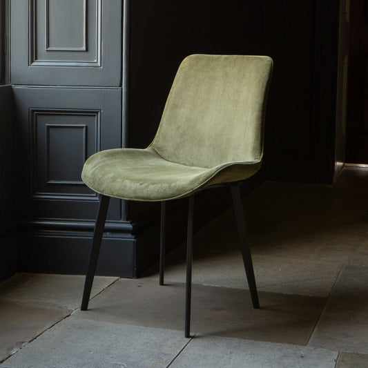 A green Eva dining chair with velvet upholstery and grey legs positioned in a room with a dark door in the background.